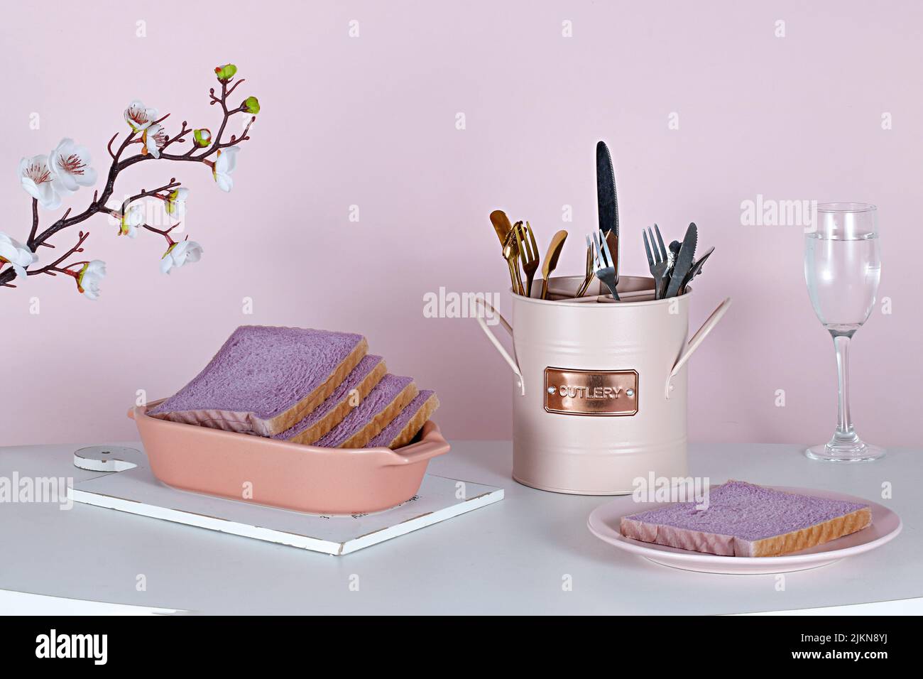 Un bel tavolo estetico con pane dolce viola, un set di posate e fiori su sfondo rosa Foto Stock