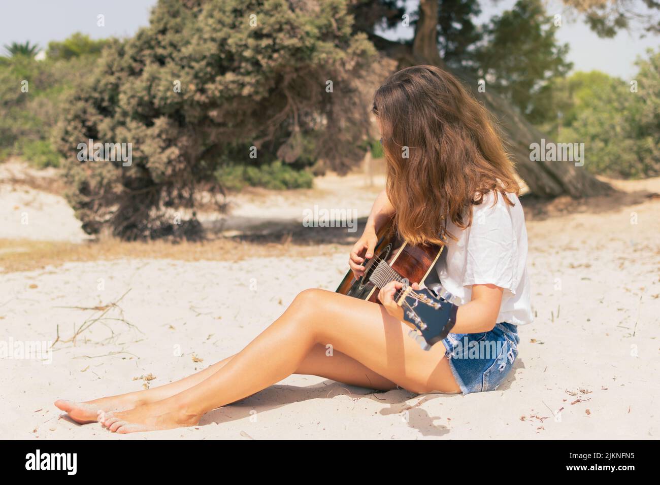 Giovane donna caucasica che suona la chitarra sulla spiaggia con gli alberi sullo sfondo. Foto Stock