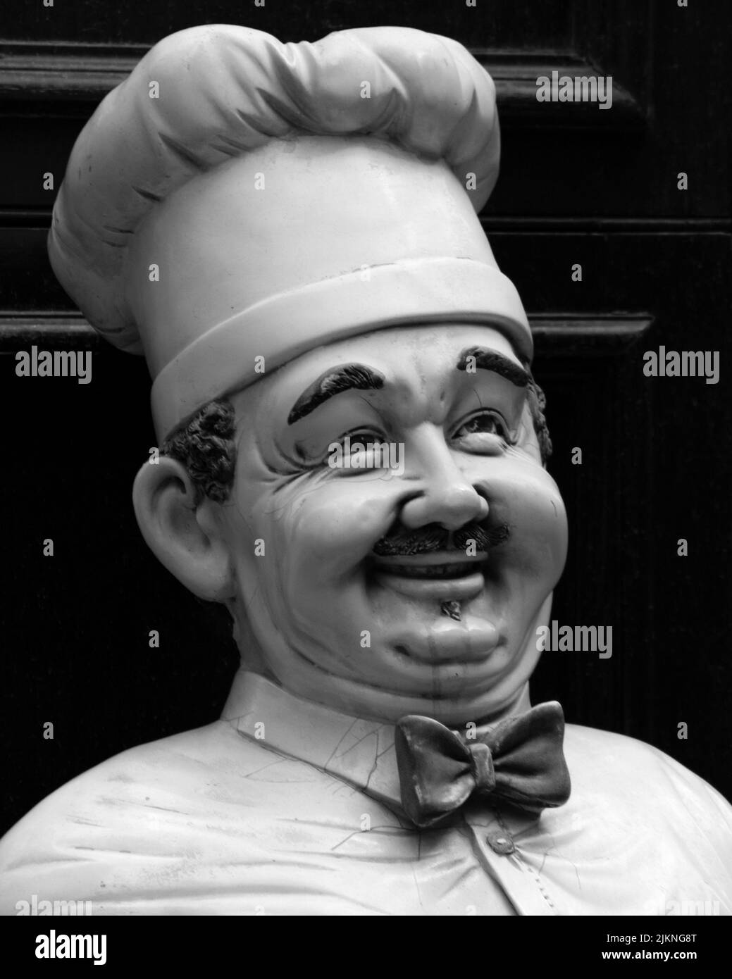 Un primo piano di una statua di un cuoco maschile vestita in uniforme con baffi e un grande sorriso in scala di grigi Foto Stock