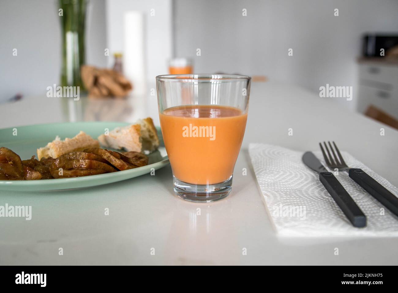Immagine di un bicchiere di Salmorejo Crema tipico piatto spagnolo con piatto verde di pane e pomodoro su un elegante tavolo da cucina bianco accanto alle posate Foto Stock