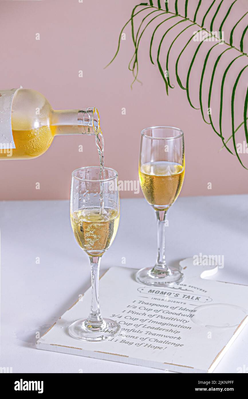 Un colpo verticale di una bottiglia di calici che versano l'alcool su un tavolo bianco e uno sfondo rosa Foto Stock