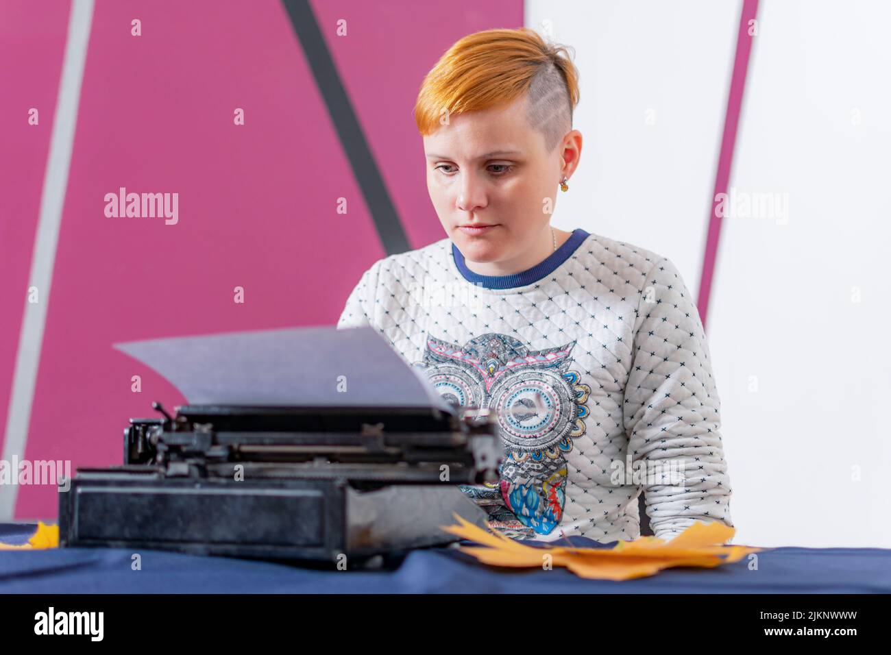 Seria ragazza giovane con i capelli rossi e stylish di acconciatura siede alla tabella e scrive il testo sulla vecchia macchina da scrivere. Foto Stock