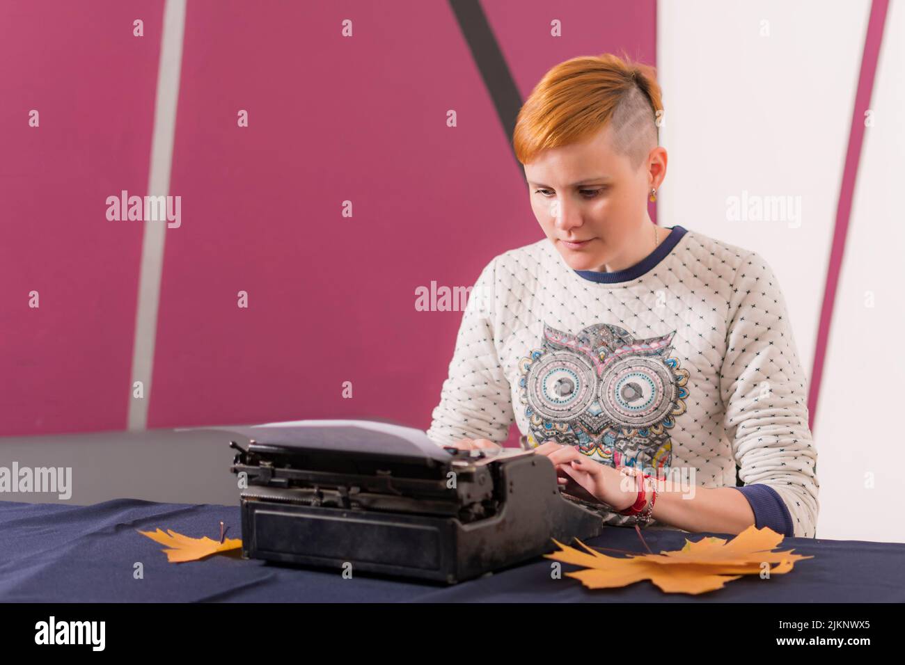 Seria ragazza giovane con i capelli rossi e stylish di acconciatura siede alla tabella e scrive il testo sulla vecchia macchina da scrivere. Foto Stock