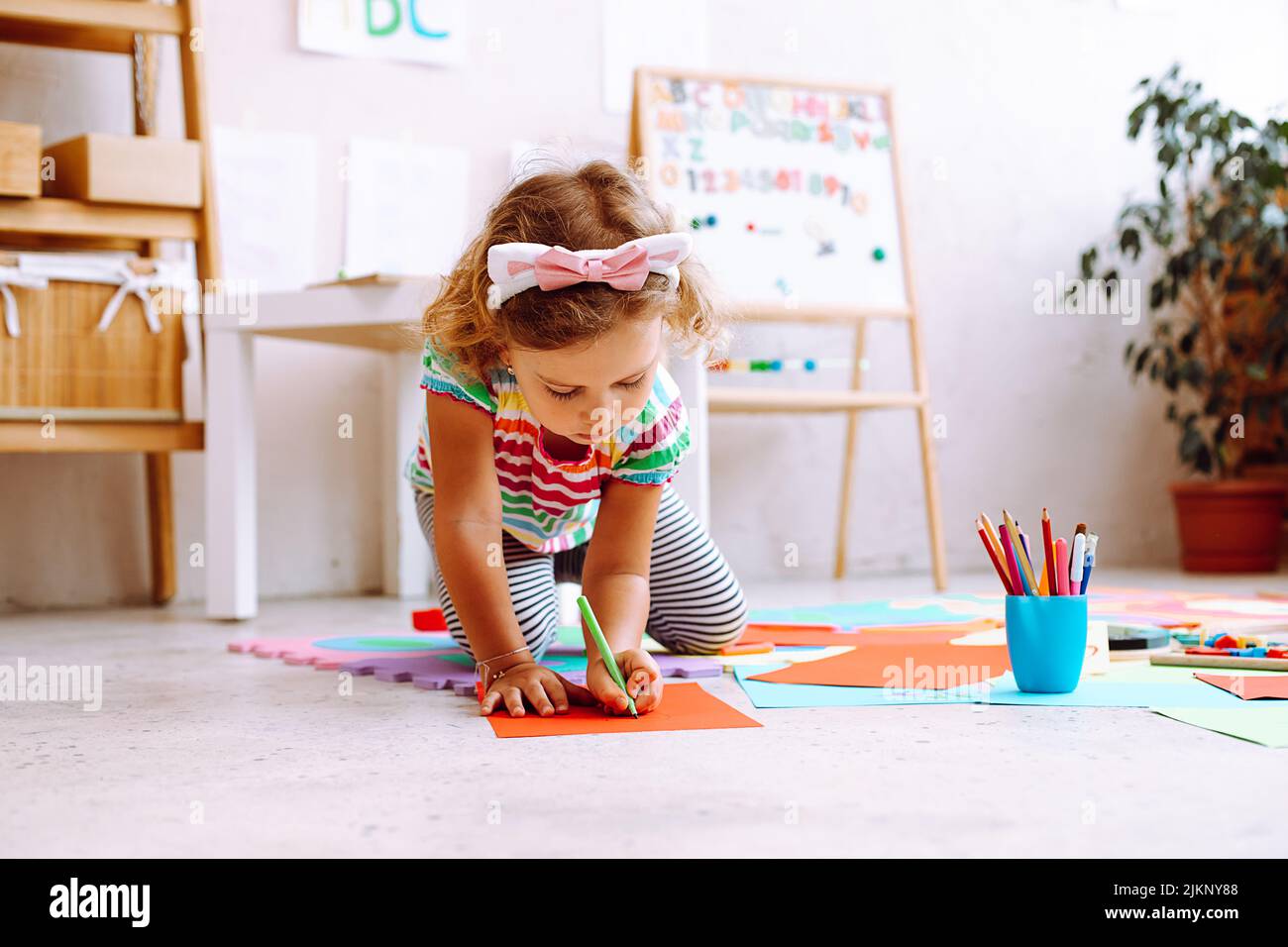 Ritratto di bambina seduta vicino tavola magnetica disegno con matita su foglio rosso di carta intorno a carte colorate. Foto Stock