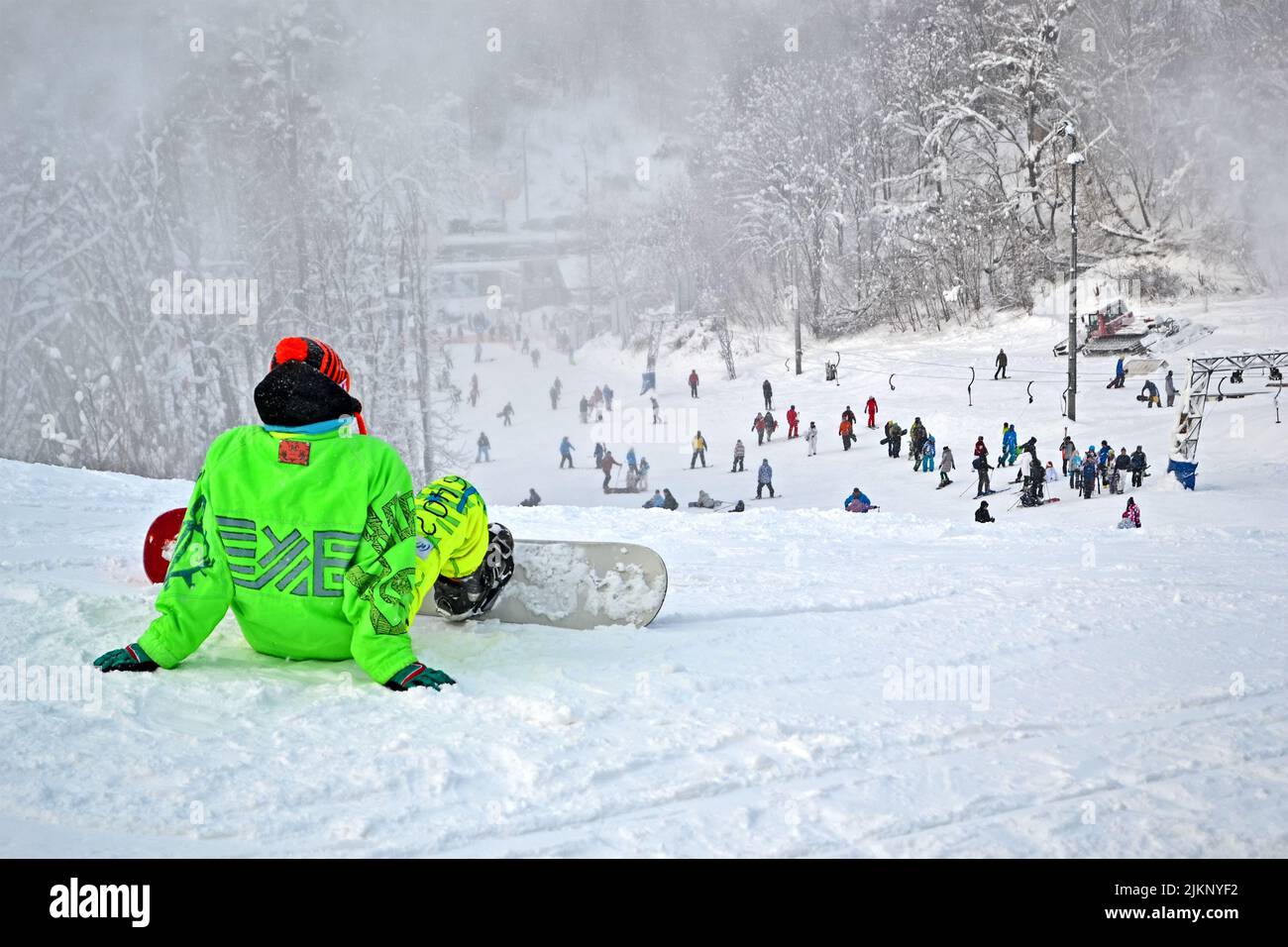 giovane sportivo in suite verde con posto a sedere in skateboard sulla neve bianca e guardare avanti, diversità sportiva Foto Stock