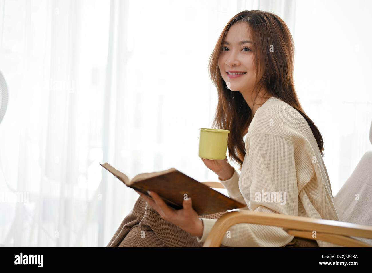 Attraente giovane donna asiatica ama leggere un libro mentre sorseggia un caffè e siede nella comoda poltrona del suo soggiorno. Foto Stock