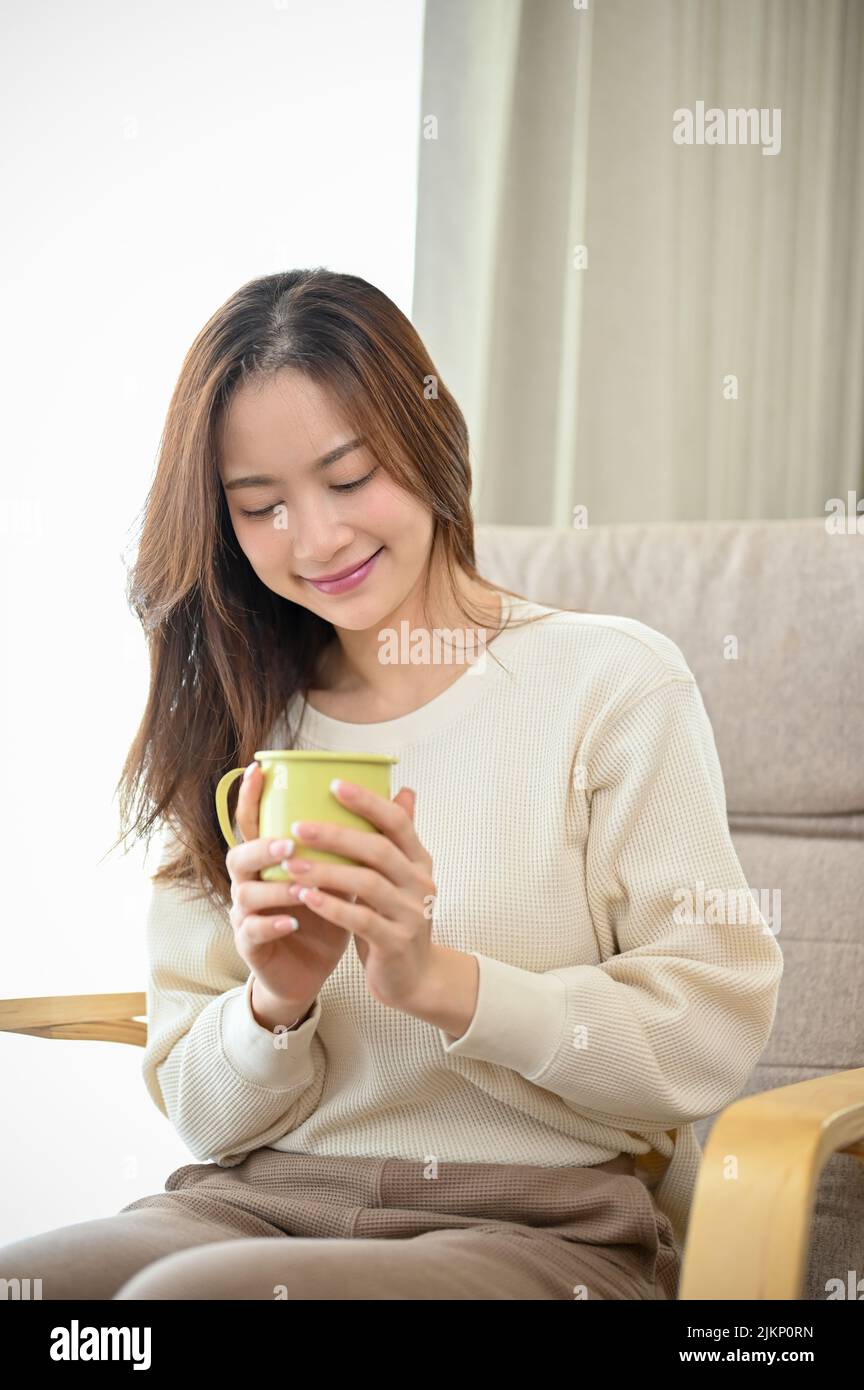 Bella e affascinante giovane donna asiatica sorseggiando caffè mentre si siede nella comoda poltrona del suo soggiorno. Foto Stock