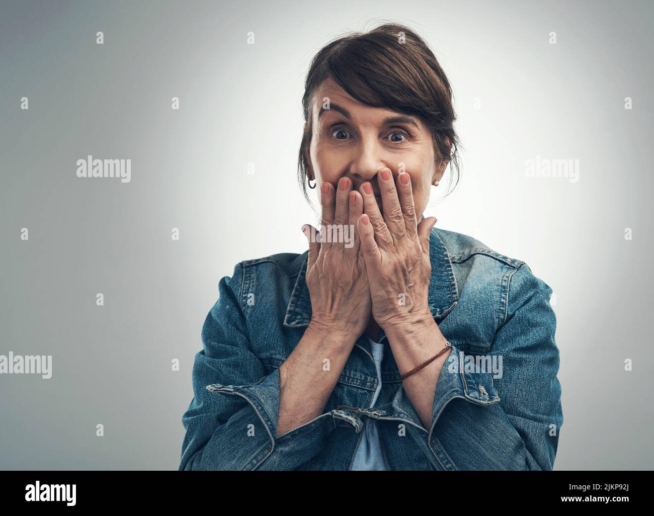 Avete sentito quello che ho sentito. Ritratto di studio di una donna anziana che copre la bocca e che guarda scioccato su uno sfondo grigio. Foto Stock