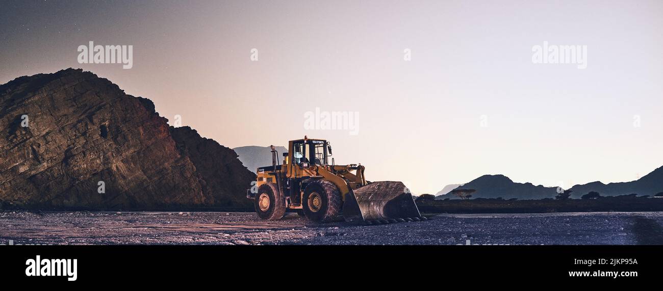 Un'immagine panoramica di un trattore che lavora nelle miniere intorno alle rocce nere al tramonto Foto Stock