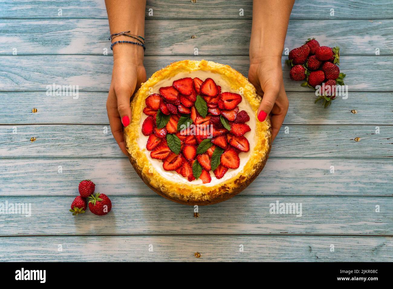 Torta di fragole fatta in casa su tavola di legno. Donna mano che tiene deliziosi cheesecake con fragole decorate con foglie di menta.Top view.Healthy Bori Foto Stock
