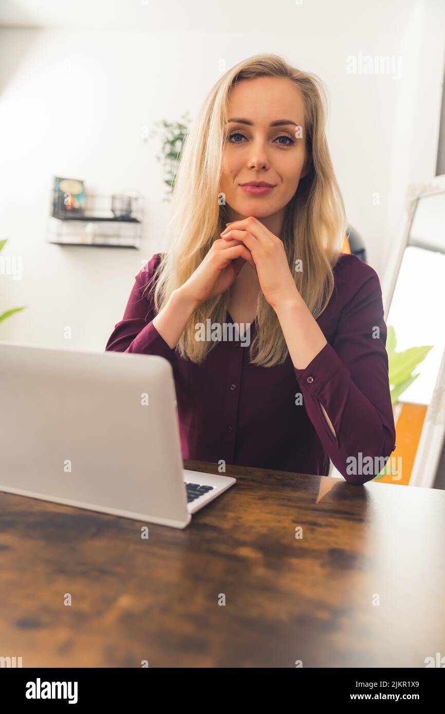 Donna caucasica con lunghi capelli biondi seduti in soggiorno con le braccia di appoggio del computer portatile sulla scrivania guardando con sicurezza nella fotocamera. Scatto verticale per interni. Foto di alta qualità Foto Stock