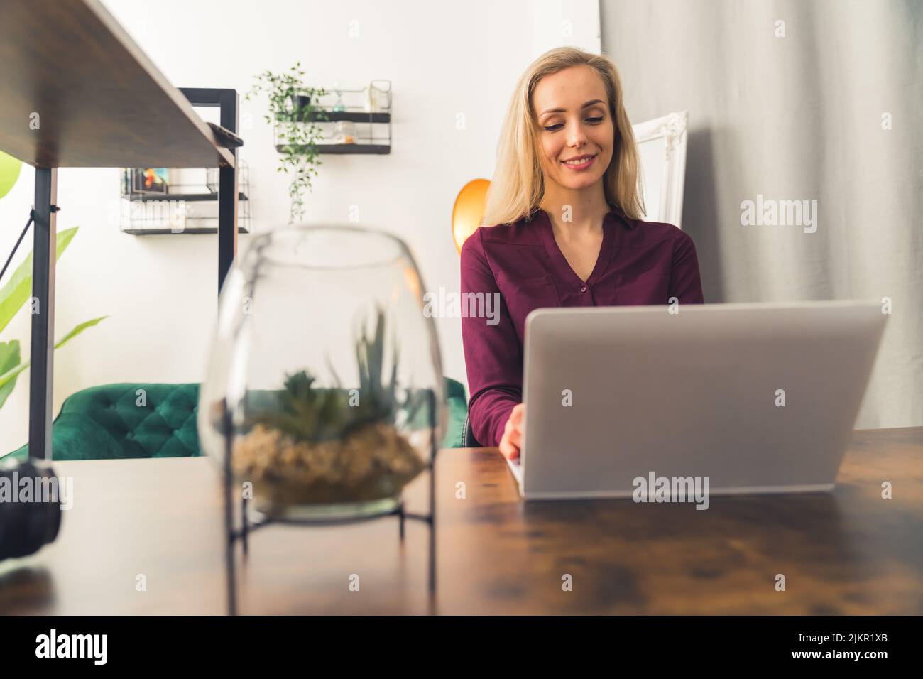 Donna bianca bionda che indossa una camicia maroon seduta sulla scrivania in soggiorno lavorando su un computer portatile e sorridendo. Scatto interno orizzontale. Foto di alta qualità Foto Stock