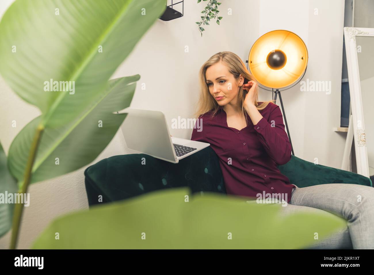 Giovane donna bionda bianca seduta sul divano verde in soggiorno tenendo e guardando il computer portatile. Scatto al coperto. Foto di alta qualità Foto Stock