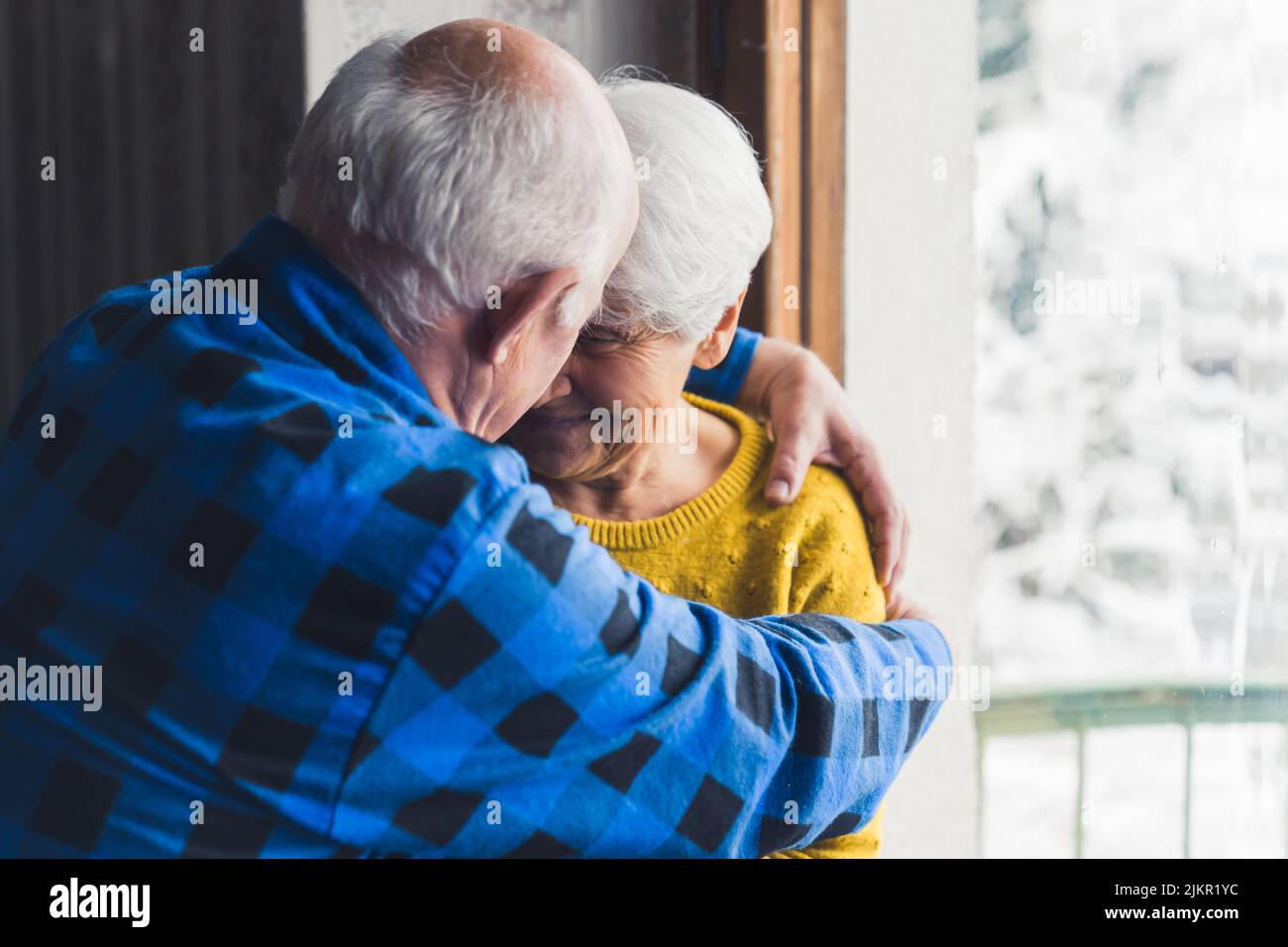 Felice amorevole marito e moglie senior sorridendo e abbracciando vicino alla finestra nel soggiorno di casa, godendo la loro vita insieme. Foto di alta qualità Foto Stock