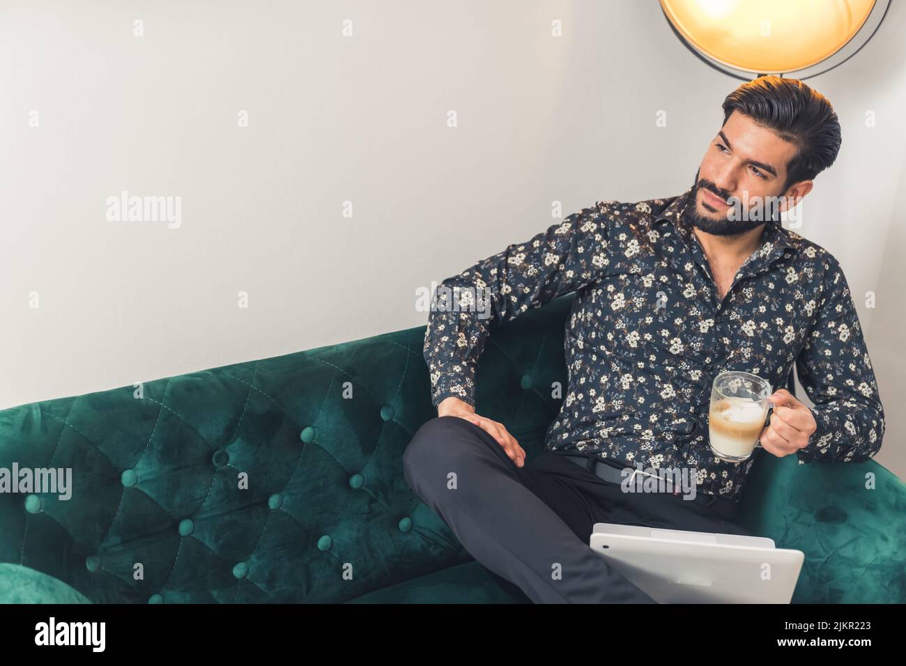 Giovane uomo ispanico che indossa una camicia seduta in soggiorno lavorando su un computer portatile facendo una pausa per bere un caffè guardando lateralmente. Scatto al coperto. Foto di alta qualità Foto Stock