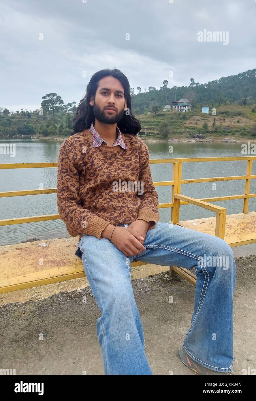 Un giovane indiano dall'aspetto buono con capelli lunghi e barba che guarda la macchina fotografica mentre si siede sulla panca accanto al lago Foto Stock