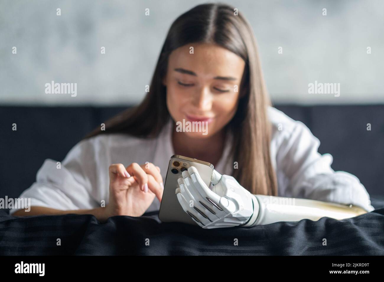 Ragazza con braccio bionico usando il telefono, leggendo i social media e chiacchierando a letto, donna con disabilità conduce la vita normale protesi articolare arto Foto Stock