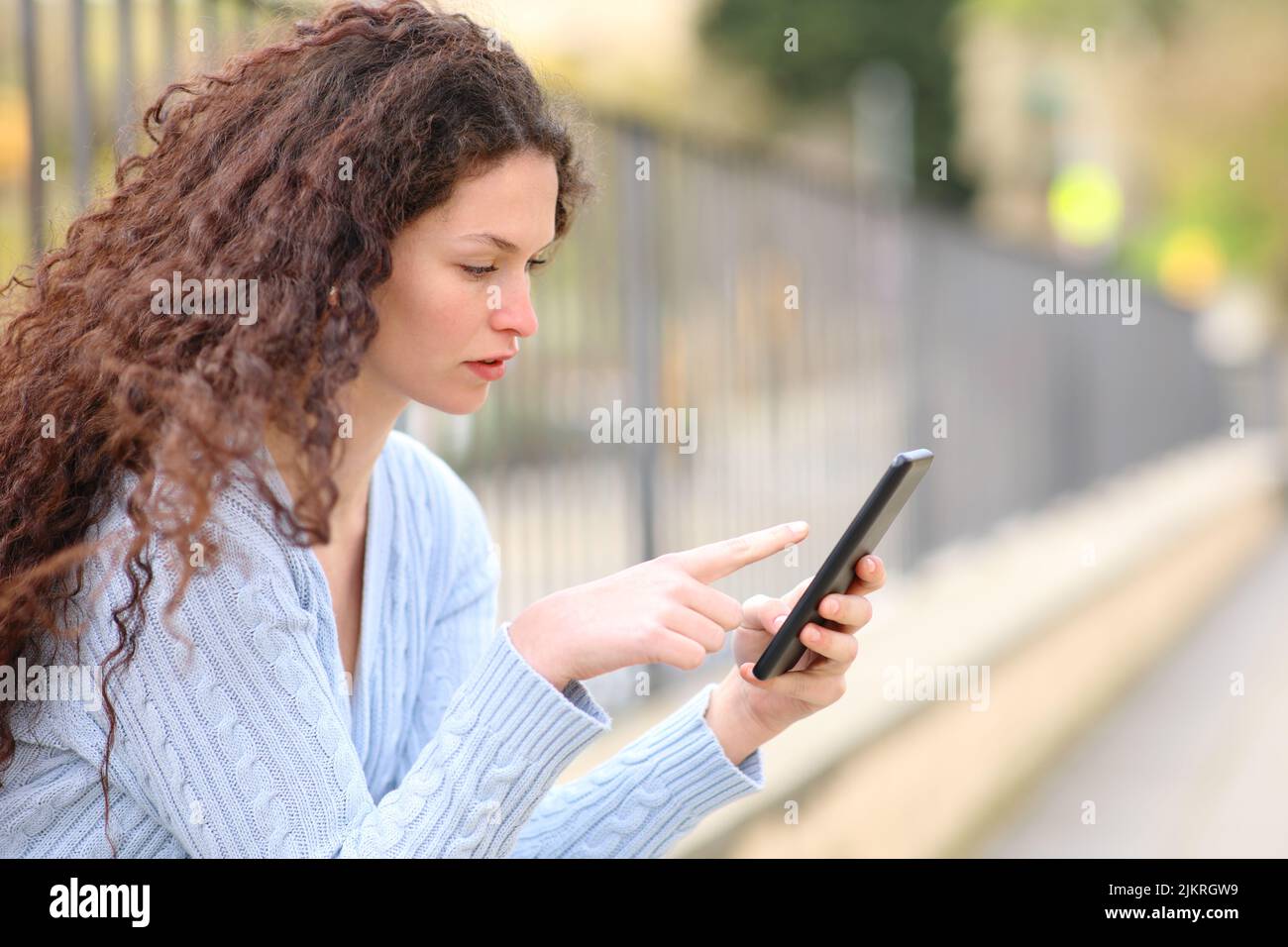 Vista laterale di una donna seria usando il telefono delle cellule che si siede nella strada Foto Stock