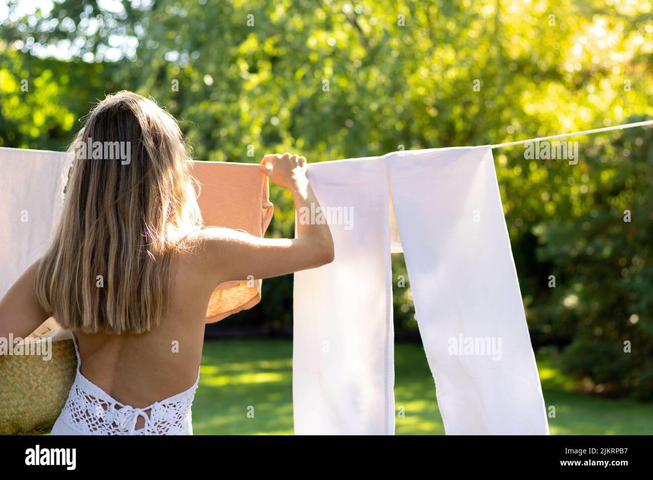 Vista posteriore della giovane bionda irriconoscibile che appende i vestiti in una linea esterna nel giardino circondato dalla natura Foto Stock
