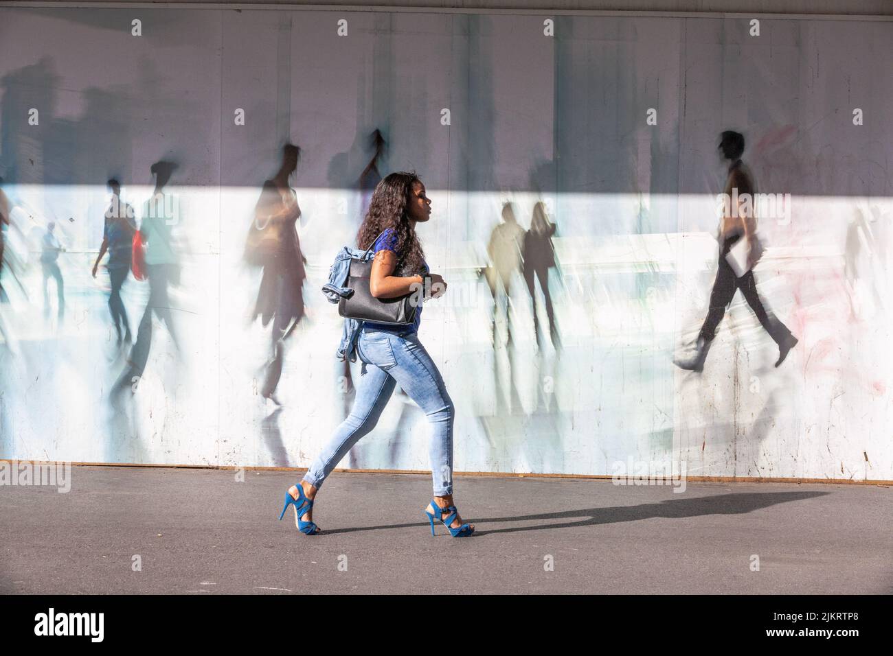 Giovane donna con tacchi alti che cammina di fronte ad una palisata decorata con figure di passanti Foto Stock
