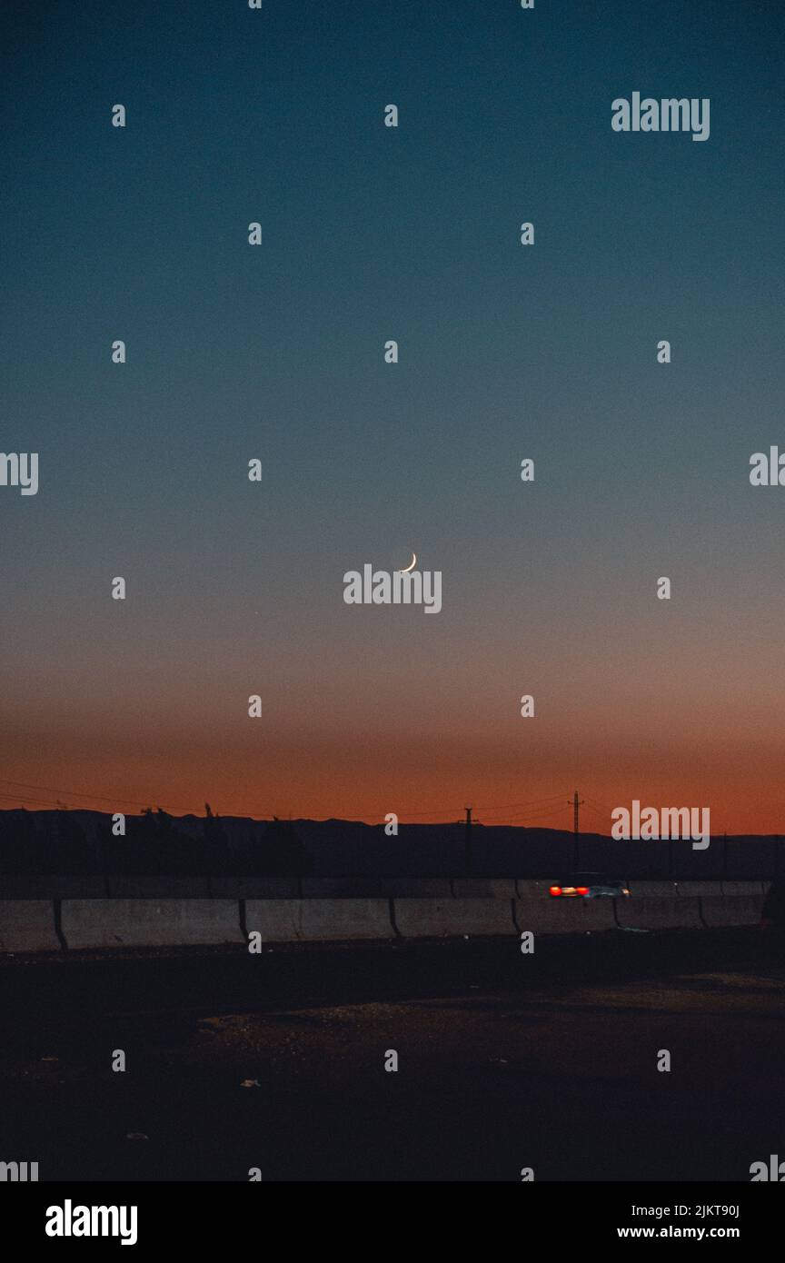 Uno scatto verticale della luna crescente nel cielo del tramonto, con un'auto che passa Foto Stock