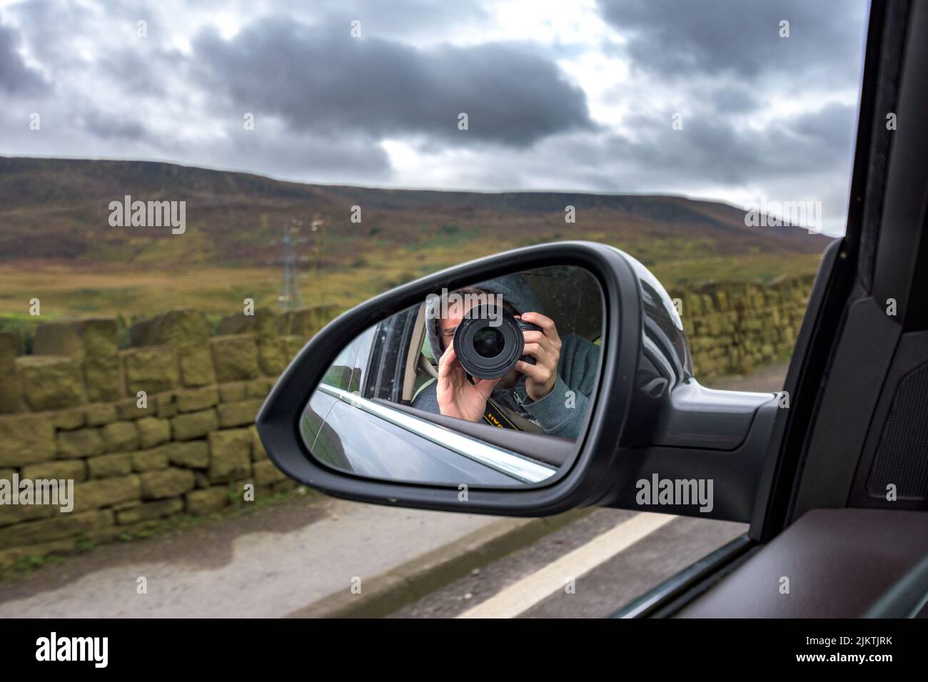 Un uomo che scatta una foto con una fotocamera nello specchio laterale di un'auto e un cielo nuvoloso sullo sfondo Foto Stock