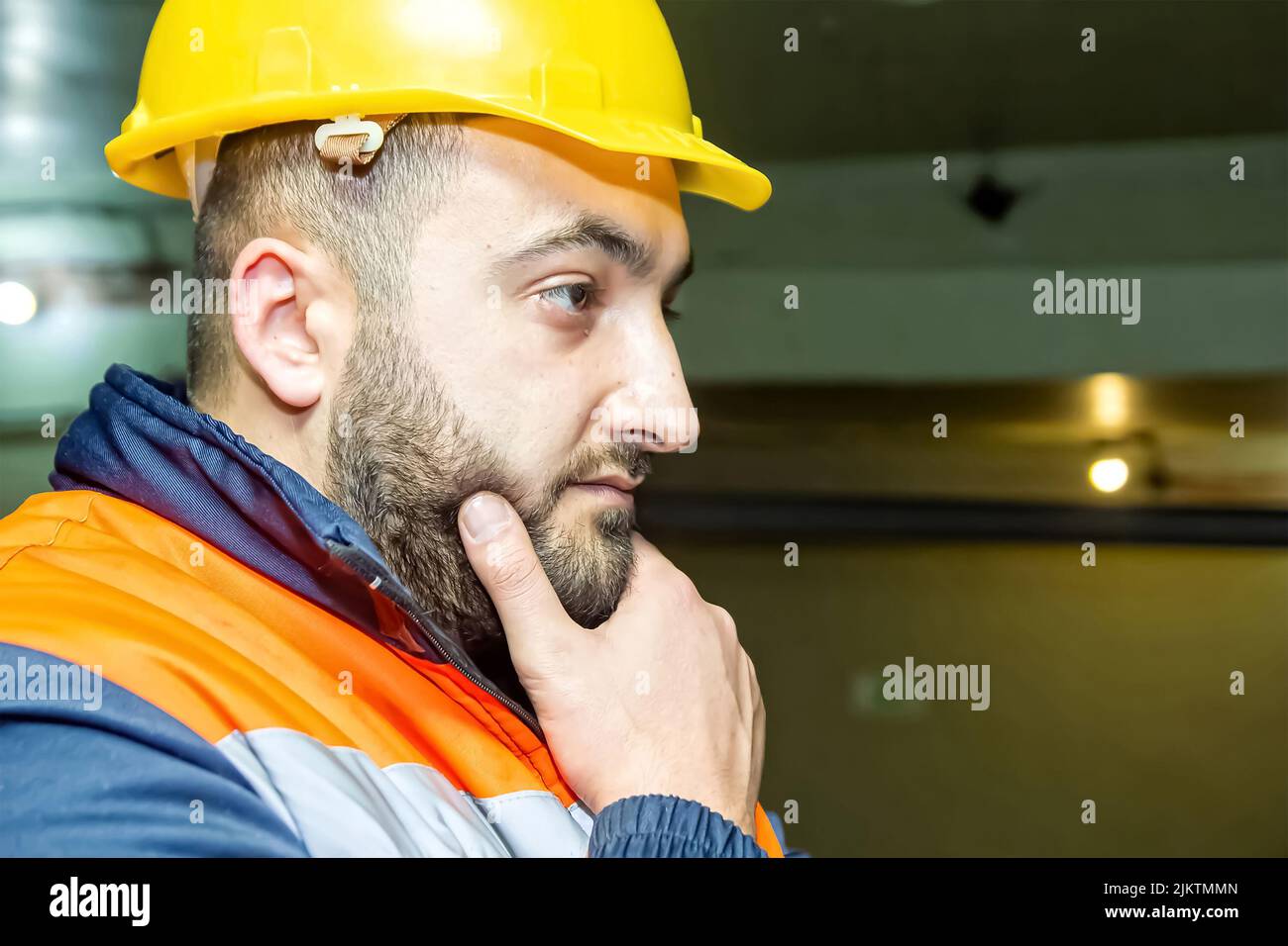 Un primo piano del volto di un uomo caucasico, che indossa un'uniforme e un casco giallo, con la mano sul mento Foto Stock