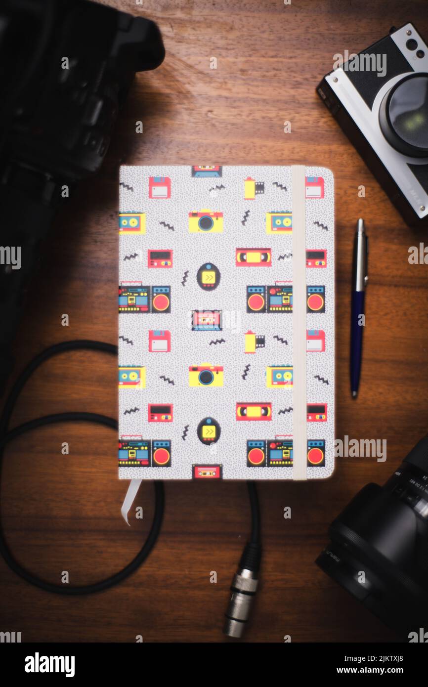 Una vista dall'alto di un notebook con icone digitali sulla copertina, una padella blu e una fotocamera su un tavolo di legno Foto Stock