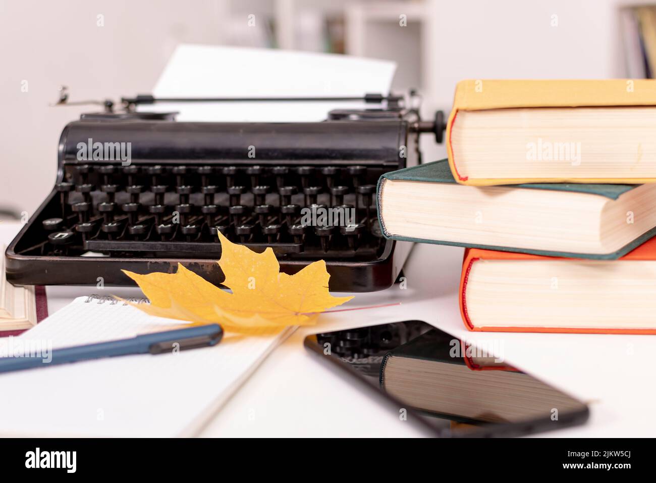 C'è una macchina da scrivere retrò su un tavolo bianco. Nelle vicinanze si trovano libri, notebook con penna, telefono cellulare e foglie di acero giallo. Luogo di lavoro dello scrittore. Foto Stock