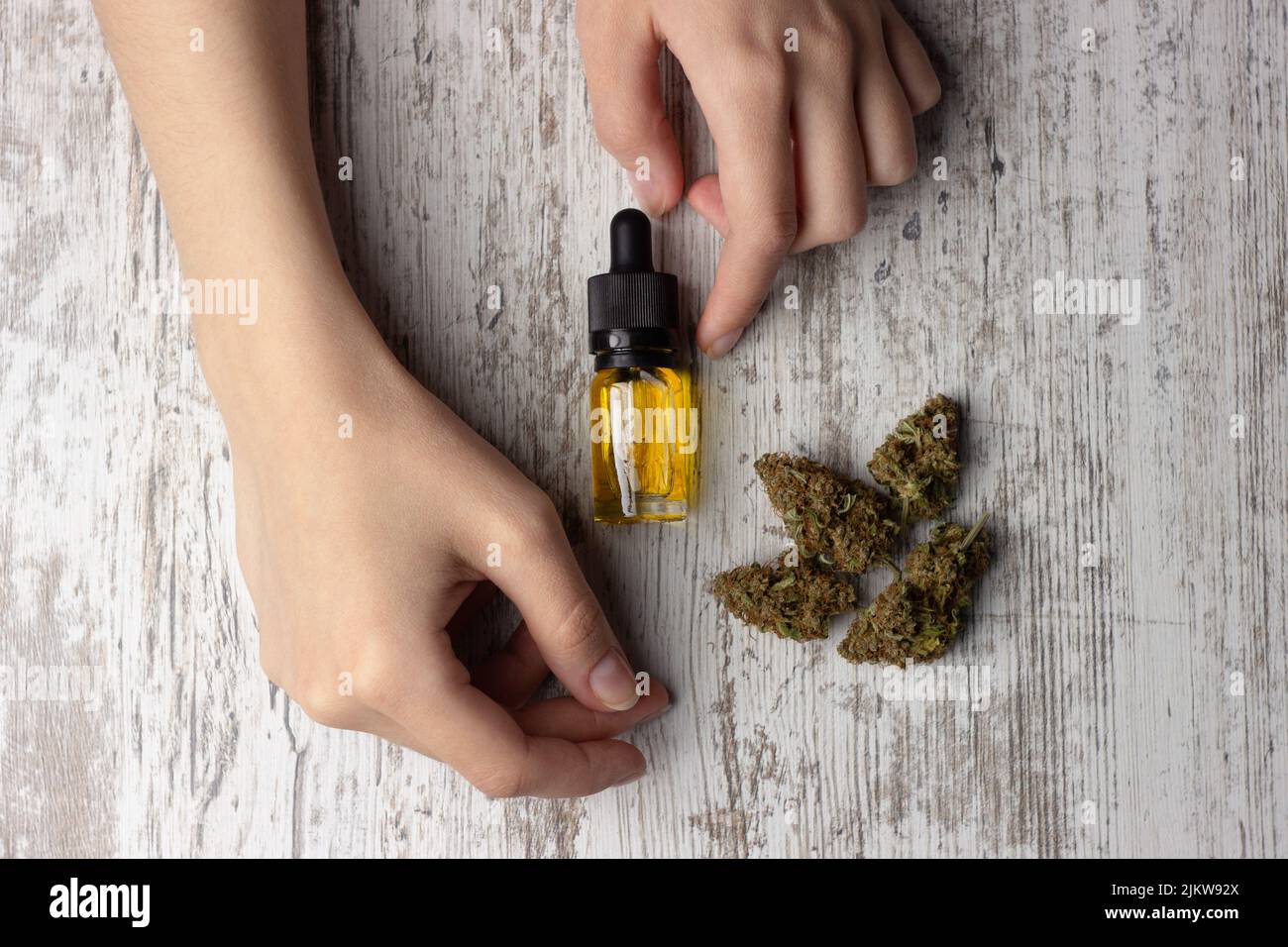 Olio cbd di cannabis in una bottiglia di vetro con una pipetta, boccioli secchi di marijuana medica si trovano nelle vicinanze. Foto con le mani femminili, su sfondo di legno Foto Stock