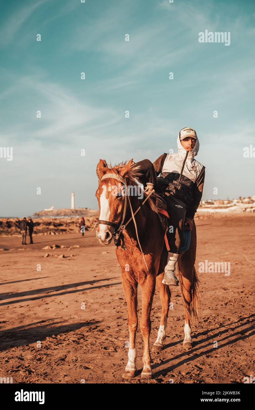 Uno scatto verticale di un ragazzo in sella a un cavallo marrone sulla spiaggia sabbiosa del marocco durante la giornata di sole Foto Stock