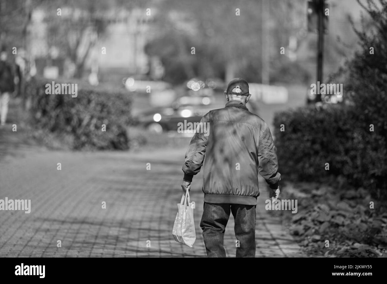 Un'immagine in scala di grigi di un vecchio uomo che tiene una borsa e cammina in una passerella nel parco in una giornata di sole Foto Stock