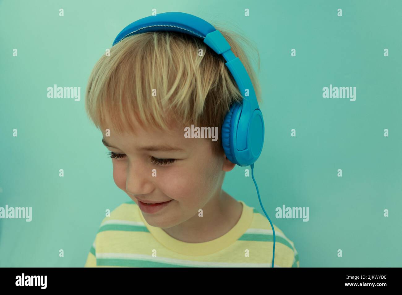 Sorridente piccolo ragazzo di 5s anni con gli occhi chiusi indossa cuffie blu, ascoltando musica. Bambino con cuffie che si diverge in classe online, tenendo vi Foto Stock