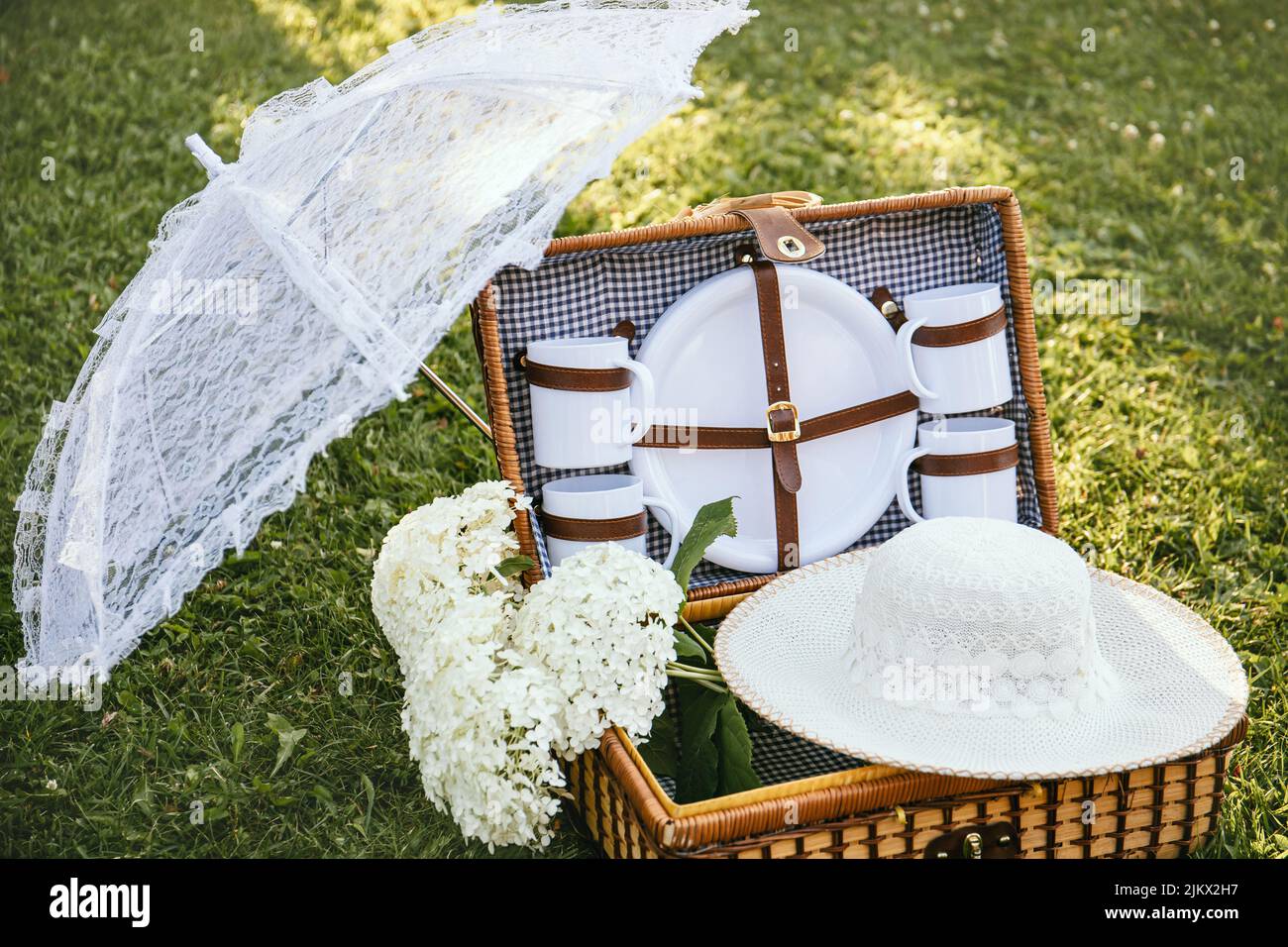 Set romantico con cestino da picnic vintage, ombrellone in pizzo e cappello estivo sul prato del giardino del parco durante le giornate di sole all'aperto. Foto Stock