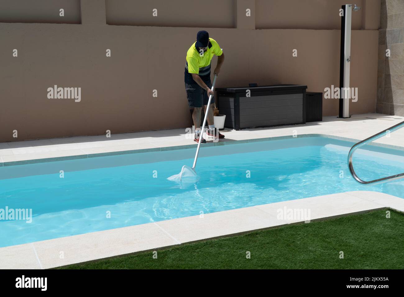 L'operatore pulisce una piscina con una trappola a farfalla, con erba artificiale e una doccia sullo sfondo. Foto Stock