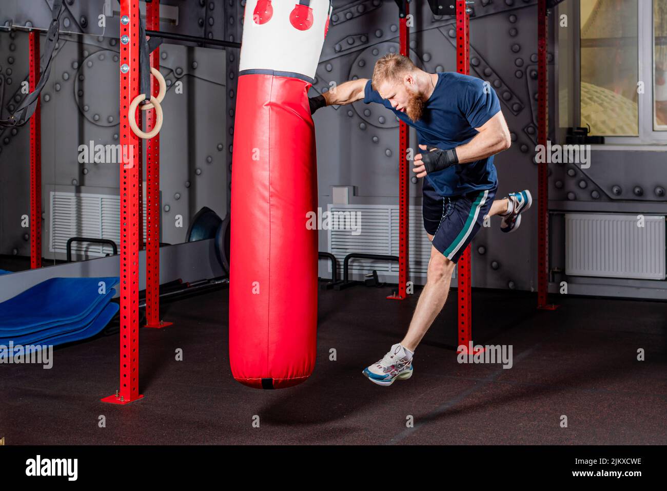 Il giovane lottatore di arti marziali miste in alto salto offre un forte pugno dritto al sacchetto rosso punzonatura. Processo di allenamento in palestra. Foto Stock