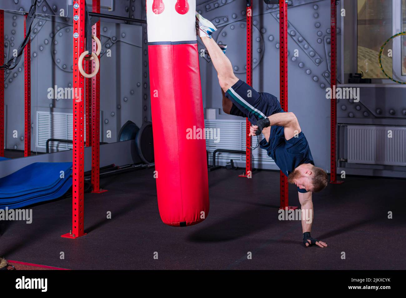 Il giovane combattente MMA in stato invertito offre un forte calcio dritto sulla pera di boxe rossa nell'area della presunta testa dell'avversario. Processo di allenamento in palestra. Foto Stock