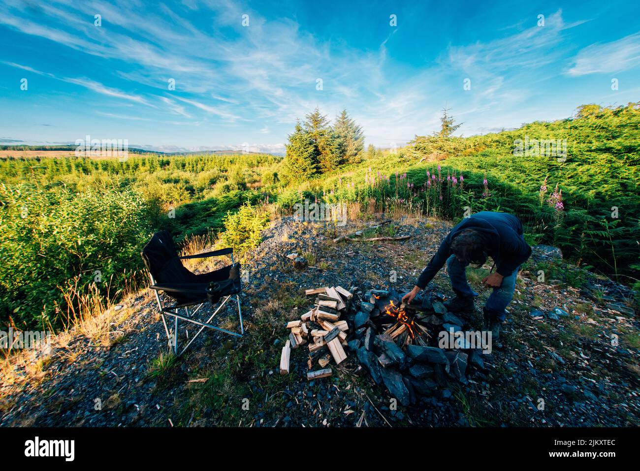Un uomo soleggiato costruisce un fuoco nel parco forestale galloway, in scozia, circondato da alberi e natura. Solitudine, fuori dai sentieri battuti, scappamento, campeggio selvaggio Foto Stock
