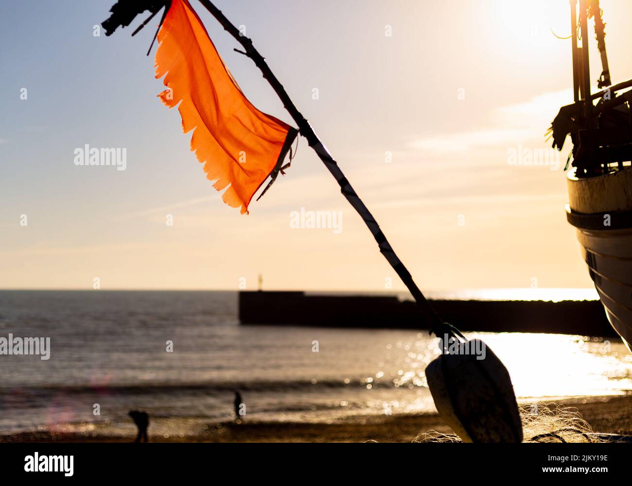 Un primo piano di una bandiera arancione su un bastone su una riva e l'acqua che riflette la luce solare Foto Stock