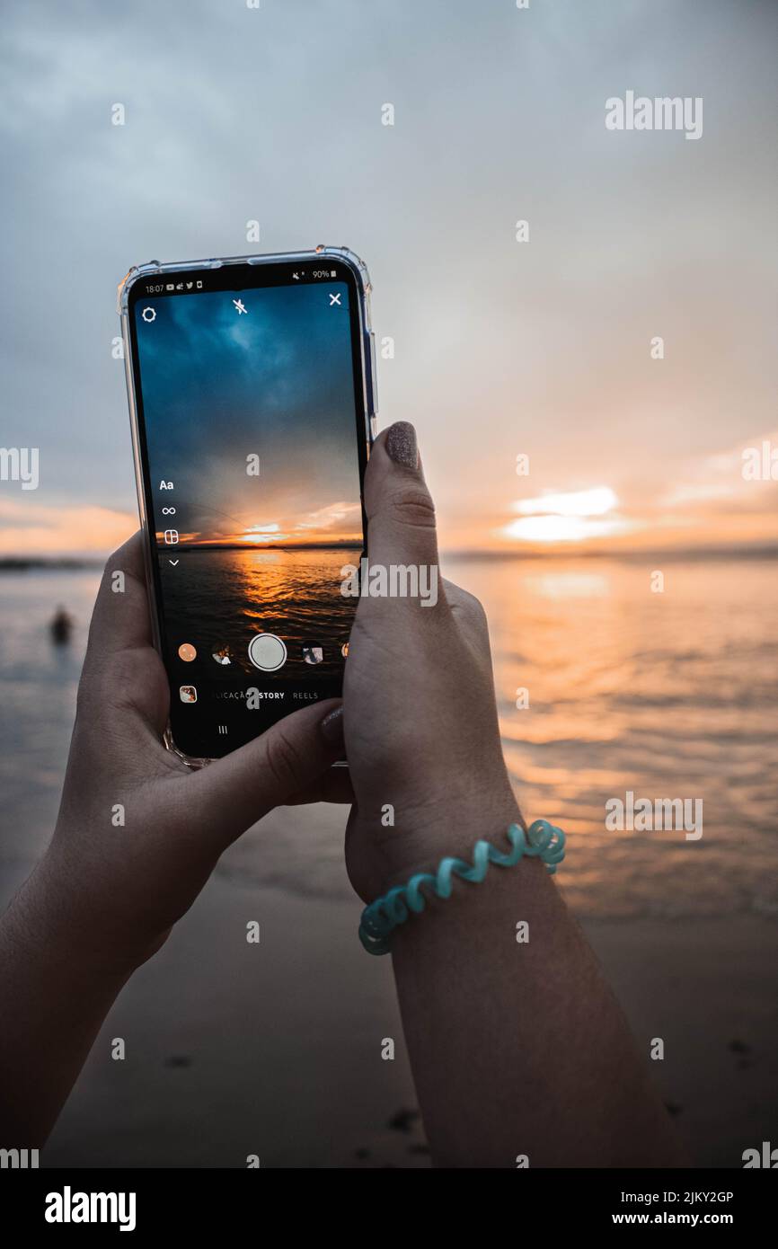 Uno scatto verticale di mani che scatta una foto di un tramonto sulla spiaggia su uno smartphone Foto Stock