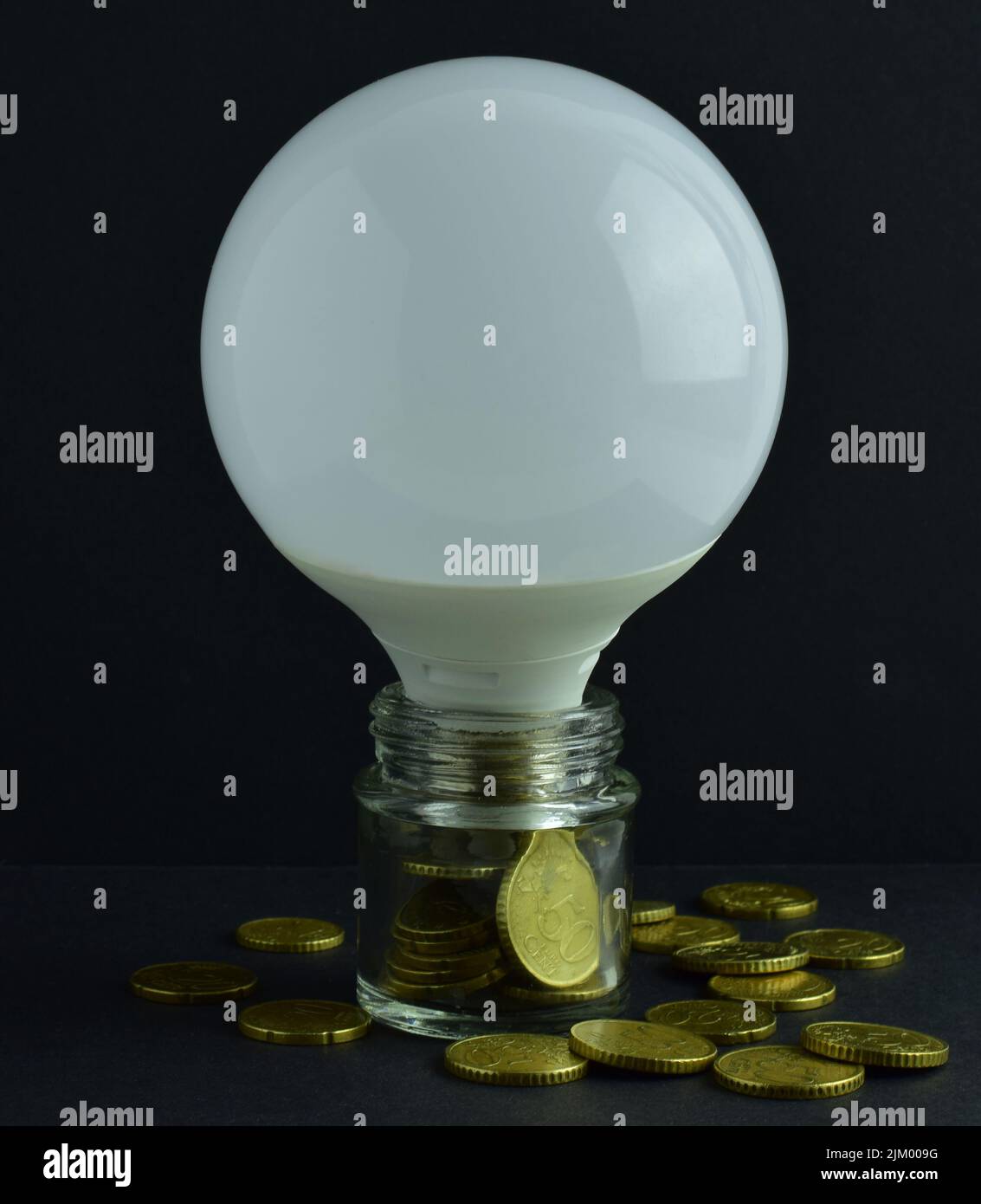 Un primo piano di una grande lampadina in un vaso di vetro con monete in euro all'interno e alcune sparse su un tavolo Foto Stock
