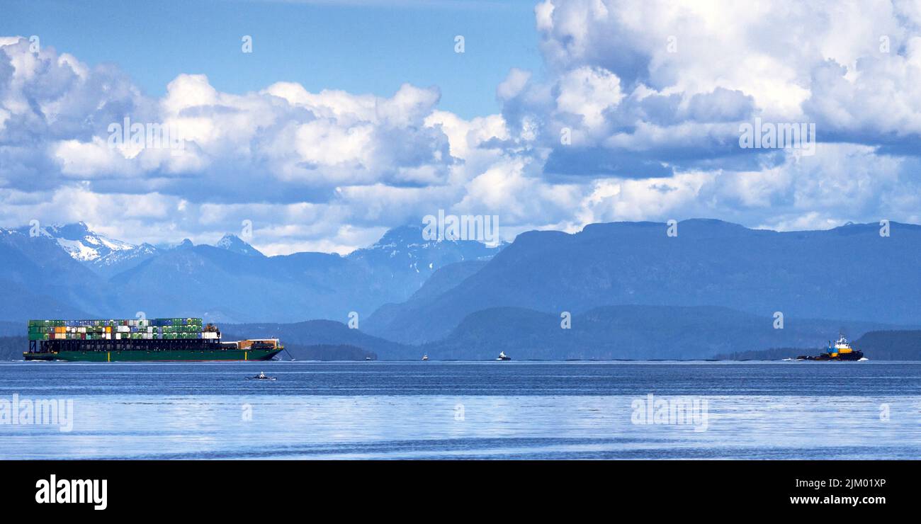 Una chiatta contenitore molto piena che viene tirata da un grande rimorchiatore giù lo stretto con uno sfondo di montagne e grandi nuvole soffici. Foto Stock