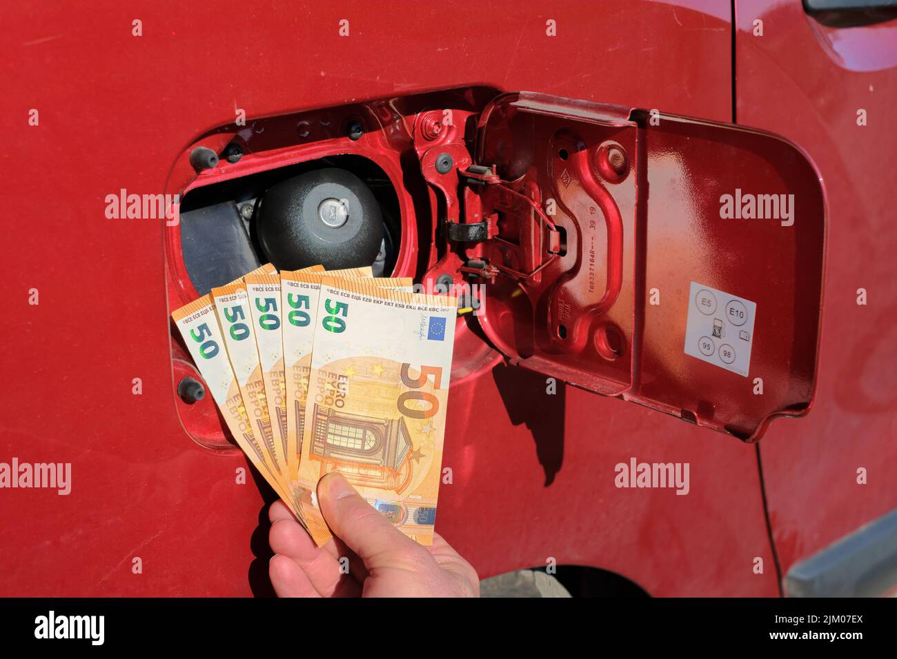 Mano che tiene un pacchetto di cinquanta banconote di euro davanti ad un serbatoio di carburante, aumento del prezzo della benzina. Foto Stock