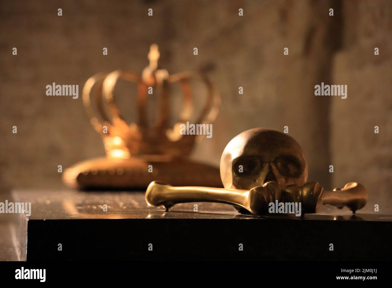 Una scultura dorata di un cranio e due ossa su un tavolo con sfocata corona del re sullo sfondo Foto Stock