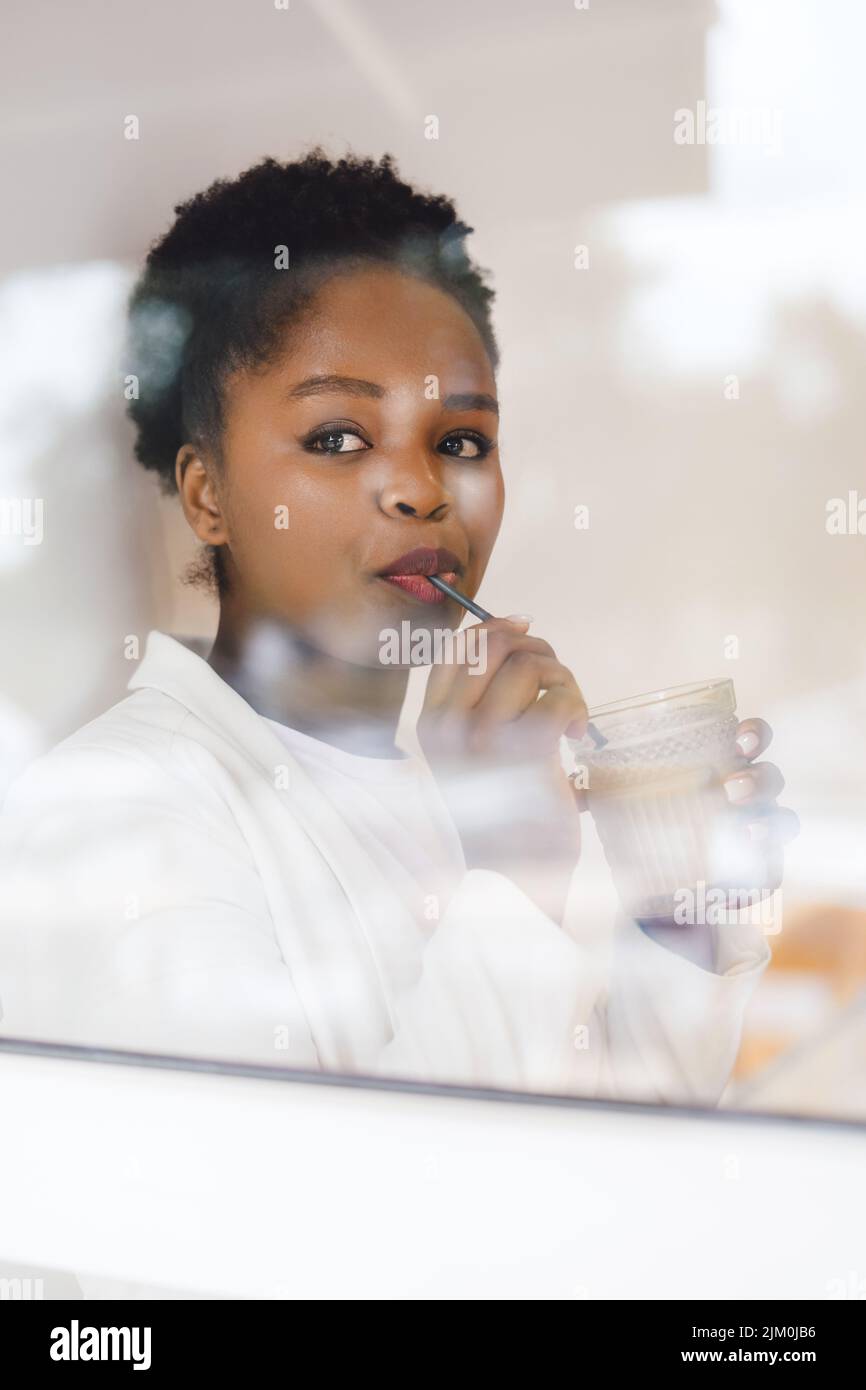 Vista esterna all'interno di un caffè dove si può vedere una donna d'affari africana bere latte con una paglia. Ritratto moderno per un design di stile. Foto Stock