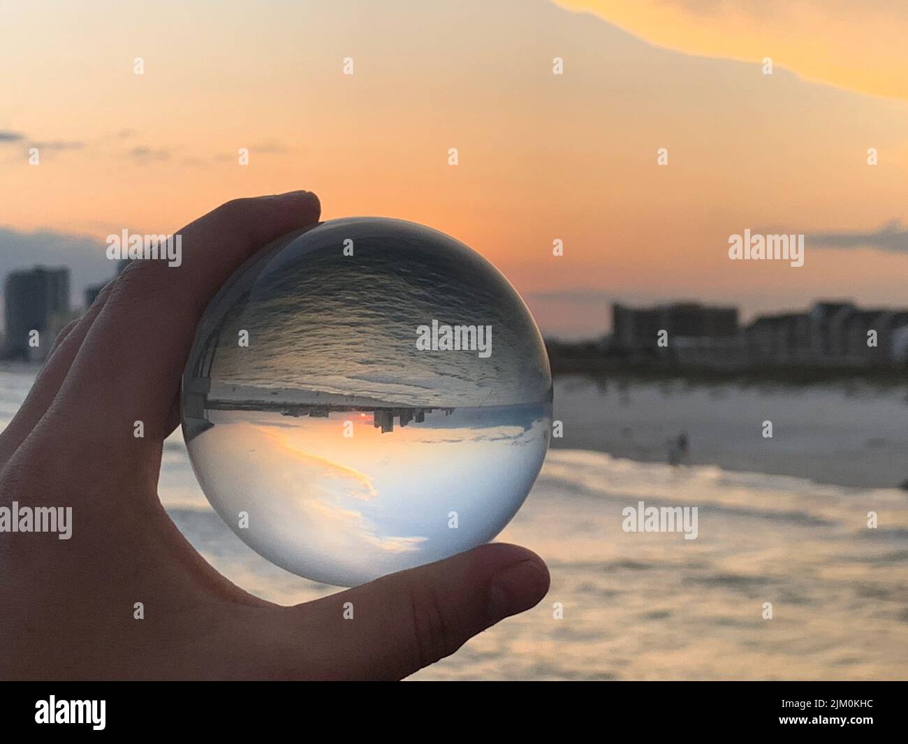 Un primo piano di una mano che tiene una palla di vetro che riflette la vista della spiaggia durante il tramonto Foto Stock