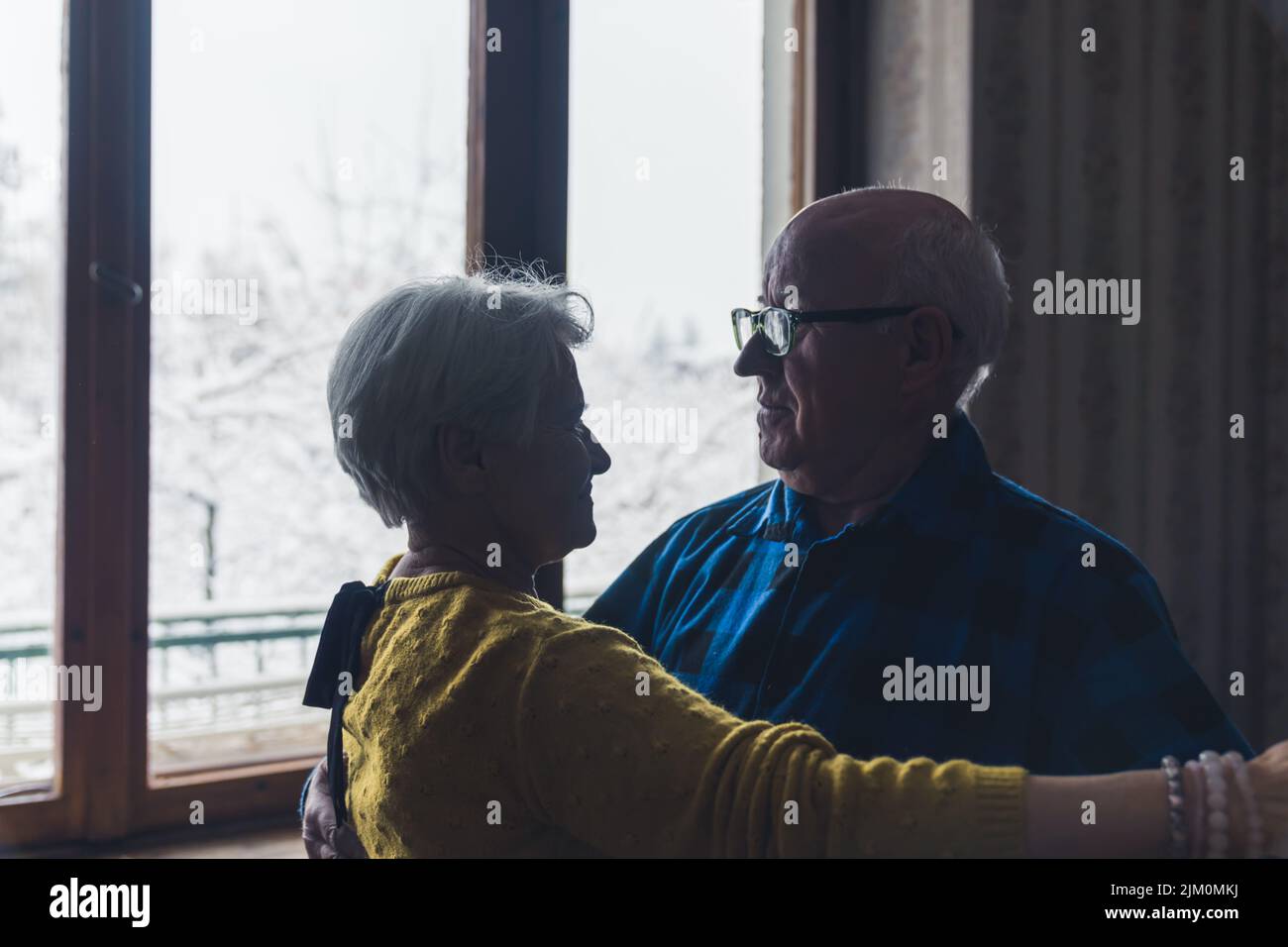 Coppia sposata senior romantica che si guarda l'un l'altro mentre balla nel soggiorno, tenendo le mani, trascorrendo il tempo insieme durante le fredde giornate invernali. Foto di alta qualità Foto Stock