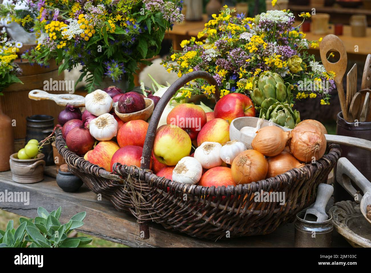 Cestino pieno di mele, cipolle, aglio e carciofi, raccolto dall'orto decorato con mazzi di fiori selvatici su un rustico tovagliolo di legno Foto Stock