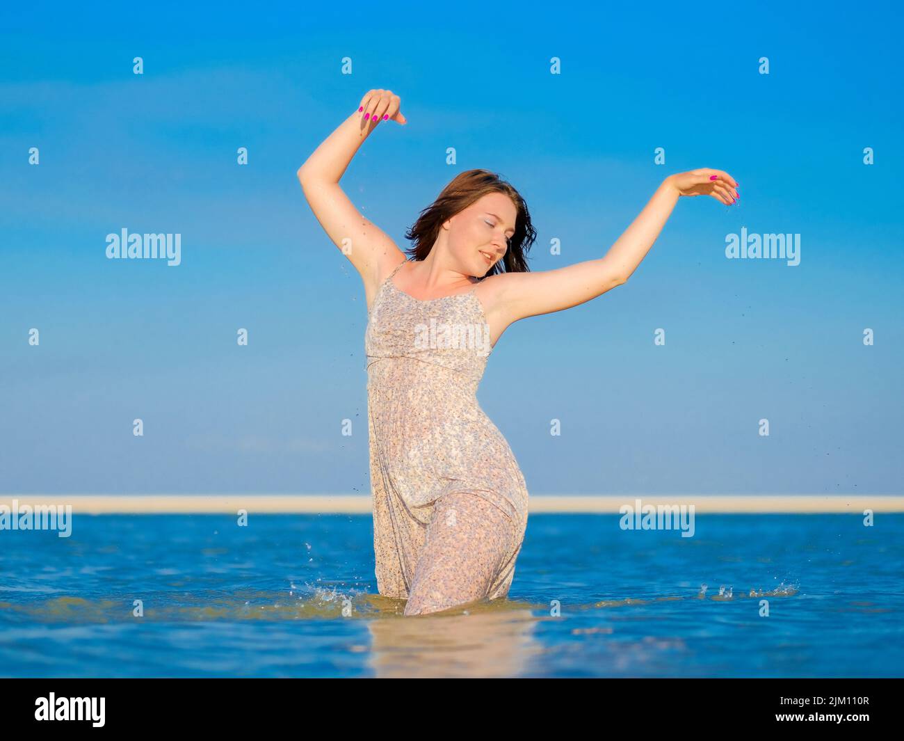Una giovane donna in una sundress bagnata gode di spruzzi d'acqua mentre sta in piedi in profondità ginocchio nel mare. Foto Stock