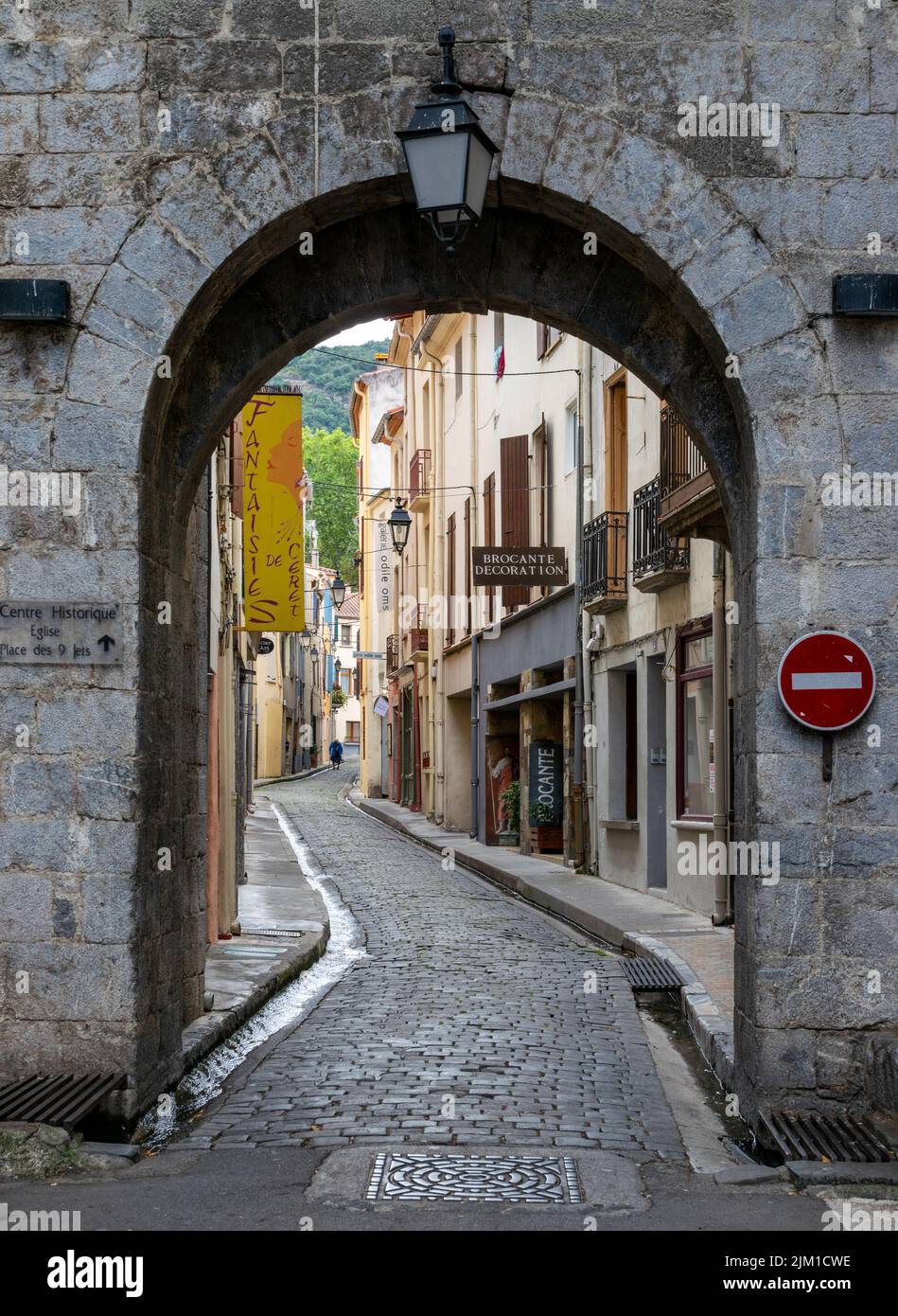 Vista di un vicolo lastricato in pietra attraverso un arco in un muro di pietra a Céret, Francia. La vista termina con le colline coperte di foresta sul retro. Foto Stock